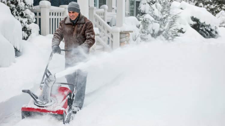 Man using snowblower to clear deep snow on driveway near residential house after heavy snowfall