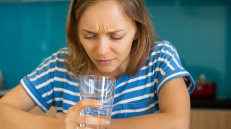 Closeup portrait of dissatisfied young beautiful woman looking into glass of water and sitting at table in kitchen