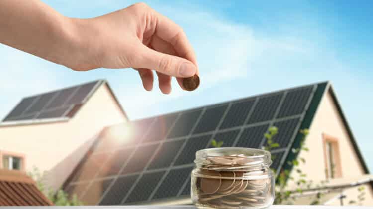Woman putting coin into jar against house with installed solar panels on roof closeup Economic benefits of renewable energy