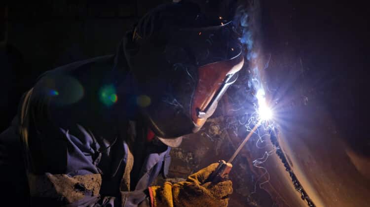 A worker in a protective mask welds a chain arc from a ditch with a pattern of abrasives against the background of a chemical factory