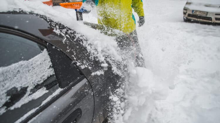 Man in rubber gloves cleans car with snow scraper on frozen asphalt Snowy and frozen vehicle after snowfall