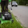 worker wearing protective clothing and gloves walks alongside the road and mows the grass with a wheeled lawnmower