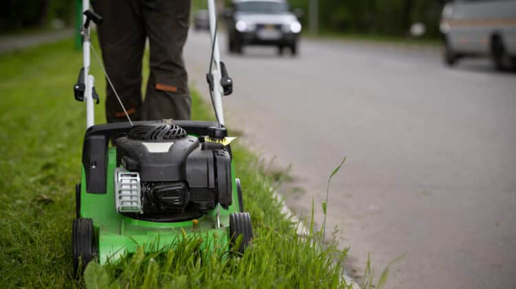 worker wearing protective clothing and gloves walks alongside the road and mows the grass with a wheeled lawnmower